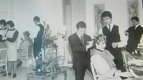 Family photograph A black and white image showing several men in suits standing above women in rollers who are sat in chairs having their hair done in an ornate room filled with grand furniture. 