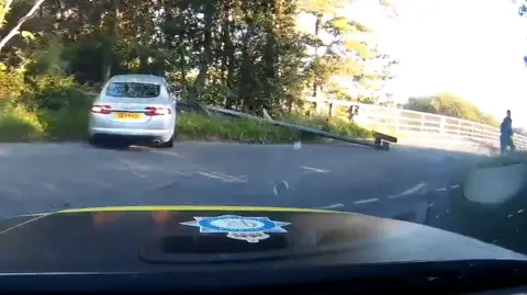 A silhouette of Daniel Lucas who is running away from a police car, which is shown in the forefront. It is a summer's day and the police car is driving on a side road which is crossing the M6 on a bridge. There is a silver Jaguar car which has crashed into a barrier in a bush.