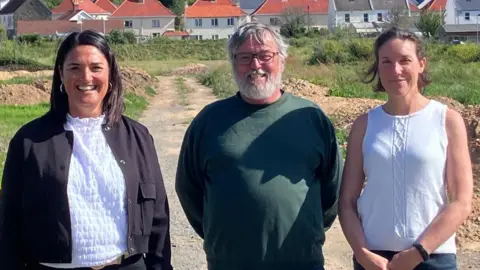 BBC Two women and a man standing in a field. The woman on the left is wearing a white top and a black jacket, the man has his hands behind his back and is wearing a green jumper and the woman on the right is wearing a sleeveless white top with her hands held in front of her.