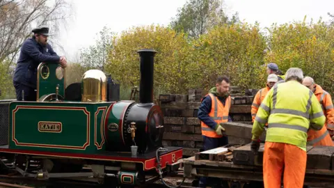 Workmen offload materials from the steam train with the name "Katie" on the side of its locomotive.