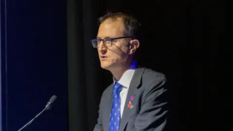 NCA Graeme Biggar is standing at a podium giving an address, he has black rimmed glasses on and brown hair. He is wearing a grey suit and a blue tie