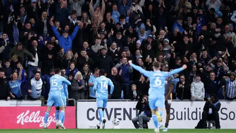 Getty Images Four Coventry players are on the pitch celebrating in light blue kits. They are running towards a stand of fans who are all on their feet and are cheering