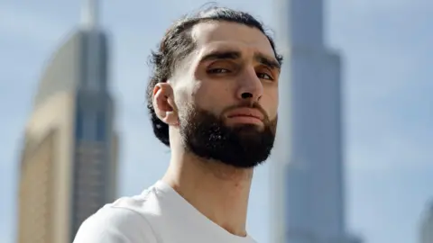 A young bearded man with long hair tied back looks down at the camera giving a tough look. Behind him, two tall skyscrapers dominate the bright blue sky.