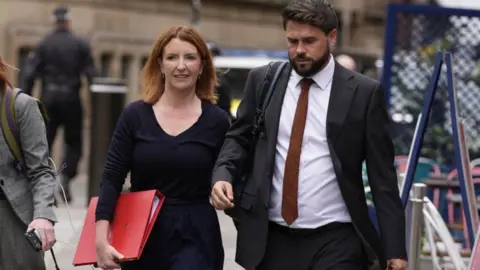 PA Media Louise Haigh is walking down the street next to an aide. She is carrying a large, scarlet government folder and wears a smart black top and black trousers. Her hair is ginger and shoulder length, and she wears it loose, along with a pair of small earrings.
