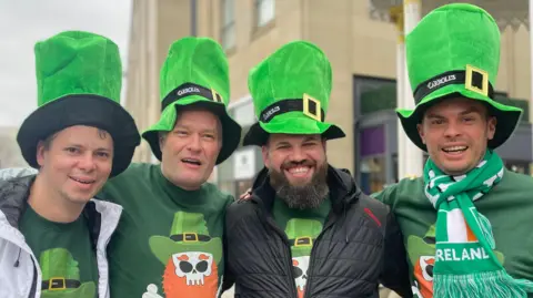 Four men are standing close together. They're wearing matching leprechaun hats and tops. 
