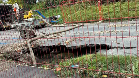Behind wire fencing, a pipe extends from a contractor's vehicle into a large hole in the road surface.