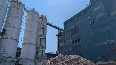 A view of the closed Unilever factory in Warrington. On the left of the image there are large white silos, and on the right there is a large blue building. In front there is a large pile of bricks from a demolished building.