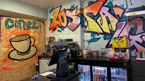 A coffee counter in the shop has a till and fridges full of canned drinks. There is a coffee machine in the background and graffiti on the walls which say 'coffee' and 'lostalgia'. 