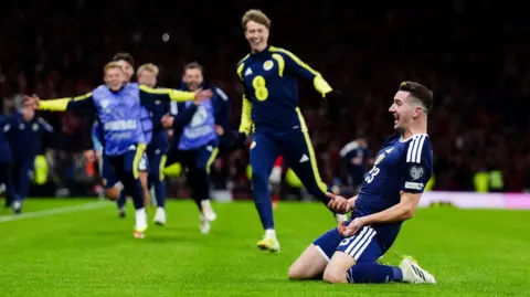 Scotland's Kenny MacLean is on his knees on the Hampden pitch as he celebrates scoring his goal against Denmark. Scotland substitutes run towards him to congratulate him.