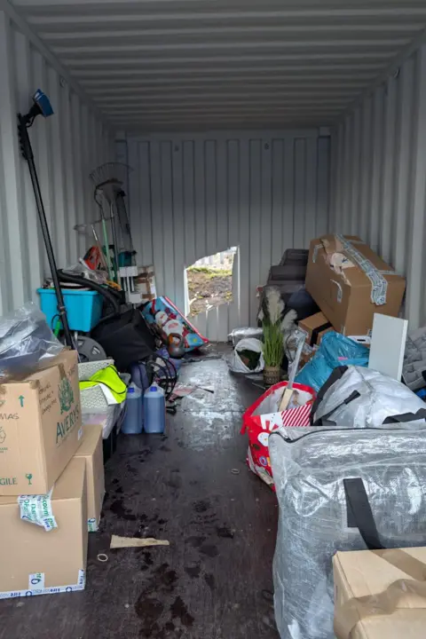 Richard Thomas Lots of household belongings in and out of boxes all stacked up and to the side of one another. A big gaping square shape cut out of the metal container, exposing the outside of the container.