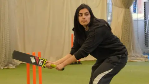 Warwickshire Cricket Foundation A girl is hunched over in playing position front of three orange plastic cricket stumps, ready to bat. She is wearing a black hoodie with black tracksuit bottoms that have a whit stripe going up the side of the leg. She is holding a black cricket bat with neon pink and yellow patterns on it. Her hands are tightly gripped around the neon yellow handle of the bat as she wears a face of concentration. She had black, straight, medium length hair which she has left down. behind her is a white sheet with netting and a green astro floor.