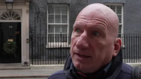 Andrew Sinclair/BBC A head and shoulders image of a man in a dark blue jacket. Behind him is the black front door of No 10 Downing Street.