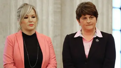 Getty Images Michelle O'Neill standing next to Arlene Foster. O'Neill has blond hair tied with a strip with a pink blazer and a black upper part, and a long silver necklace. She carries makeup with pink lipstick. Foster has short and brown hair and wears a pink striped shirt with a black blazer.