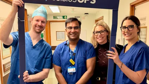 A group of medical professionals wearing blue scrubs are standing in a hospital hallway smiling at the camera.