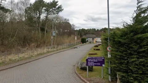 A paved entrance way to a council run caravan park with trees down the sides and a 5mph speed limit sign