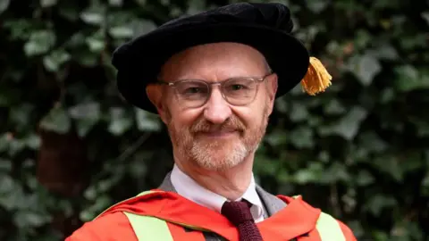 University of Leeds Mark Gatiss in a red and green graduation gown and a black velvet graduation tam hat.