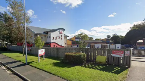 Busy Bees Nursery behind a grassy area and brown fence. It is over multiple floors and has red and white paint on the walls. 