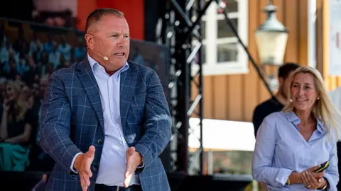 News Room : Tight vote in Norway to decide whether to stick with Labour or turn right 2 Bloomberg via Getty Images A man in a checked-blue jacket and light blue shirt stretches his hands out as he talks, while a woman looks on
