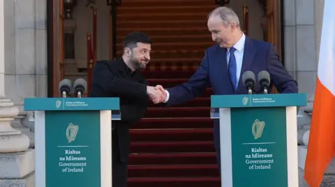 PA Media Ireland's Taoiseach (Prime Minister) Micheal Martin shakes hands with Ukrainian President Volodymyr Zelensky at Government buildings during an Irish State visit, in Dublin, Ireland. Martin (left) is wearing a navy suit. Zelensky (Right) is dressed all in black. They are standing at Government of Ireland microphone podiums.