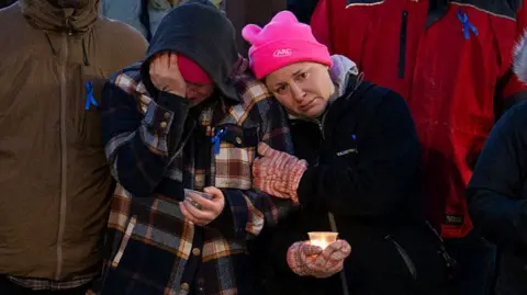 Two people are seen at a vigil, both holding candles in their hands. One is leaning on the other to console them as they cry.