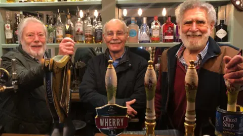 Three men standing behind a bar, with a row of beer pumps in front of them. The man on the left has white hair and a white beard. He is wearing a dark green outdoor jacket. The man in the middle is bald, with a white moustache and is wearing glasses. He is also wearing a dark blue outdoor coat. The man on the right has curly white hair and a white beard and moustache. He is wearing a tan and black gilet over a red jumper. There is a row of spirit bottles on a shelf behind them.