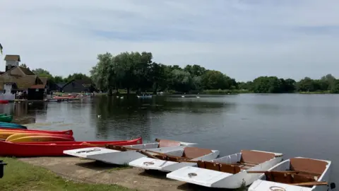 Boats are on the edge of a lake, buildings can be seen on the left edge of the lake with trees on the far side. Some bird can be seen swimming on the water.