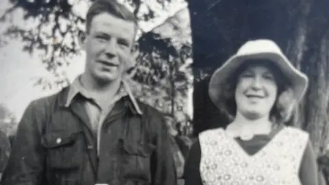 Walter Renwick A black and white photograph of two members of Walter Renwick's family taken perhaps in the 1940s or 50s. The man on the left is wearing overalls and a shirt, the woman on the right has a pinafore and sunhat. There is a large tree visible behind them.