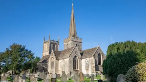 The North Wessex Way A medieval church on a sunny day. The church has a square tower and a spire. Around the church are many gravestones. 