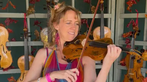 Andy Parish A lady with blonde hair done up in a bun with a small flower in, she is playing a violin and is wearing a pink top with thin straps and multi-coloured chiffon scarf around her neck, behind her are several violins hanging up.