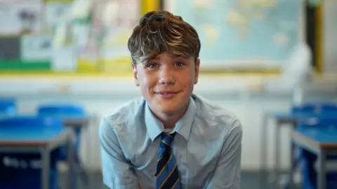 Gemma Laister / BBC A young school pupil, Ethan, smiles as the camera. He is wearing a school uniform - black, yellow and blue striped tie and pale blue shirt - and sitting in a classroom. 