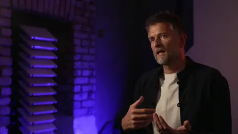 Architect Sean Hill, wearing a white T-shirt beneath a black shirt sits in a purple lit room