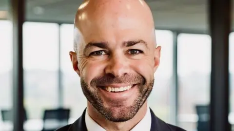 Michael Becker Head and shoulders shot of Michael Becker pictured inside an office building. He has a shaved head and a short dark beard. He is smiling at the camera. He is wearing a dark suit jacket and white shirt.