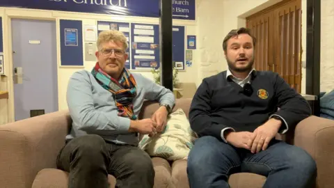 Choir members Dominic Fawcett (left) & Lloyd Evans (right) sitting on a couch during an interview. Mr Evans is talking to the interviewer. Mr Fawcett is looking at the camera. There is a cushion on the couch. There is a notice board behind them.