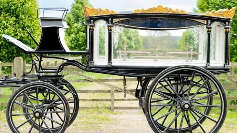 Duggleby Stephenson Black Victorian London horse-drawn hearse parked in front of a gate.