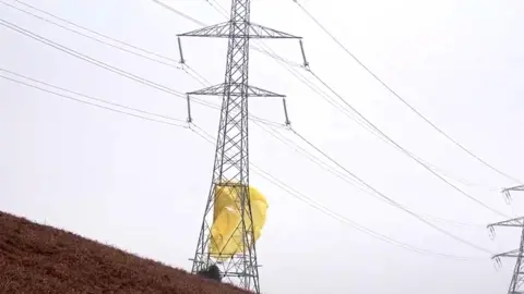 A large electricity pylon with yellow material resembling a parachute wrapped around the bottom of it.