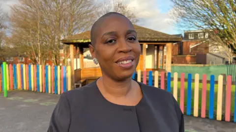 Headteacher Tracey Adams is a woman in her 50s, smiling at the camera, in a smart black coat and silver circular drop earrings. She is stood infront of the school playground, which has a hopscotch in the background, a rainbow colours wooden gate and sheltered area.