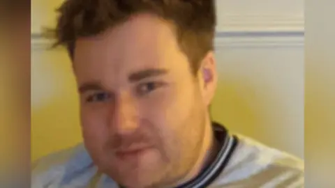 South Yorkshire Police Jamie Kelly looks at the camera. He has short, spike brown hair. He is wearing a grey top with a blue and white crew neck collar.