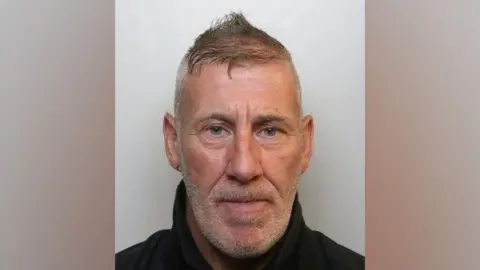 Avon & Somerset Police A custody picture of Wayne Murray. He has grey hair, short on the sides and long on top, and short grey facial hair. He wears an earring in one ear and a dark fleece, and is looking directly into the camera with a neutral expression. 