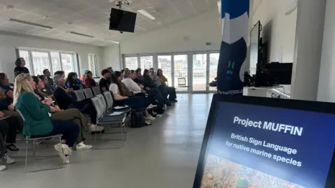 A group of people sat looking at a screen at a conference. There is a sign with the words 'Project MUFFIN' to the right with the words below 'British Sign Language for native marine species'.
