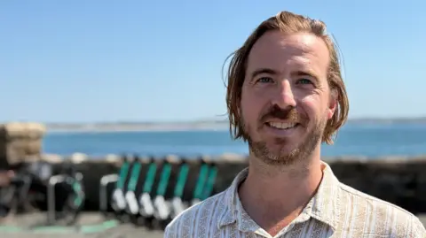 Phil Ellis - a man with blond hair and a beard. He wears a white and gold patterned shirt. He is standing on the prom in Penzance with a row of e-scooters and the sea behind him.