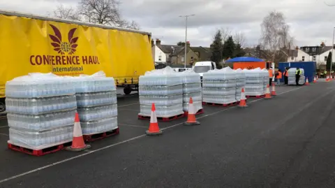 Jack Fiehn/BBC Crates of bottled water are lined up in front of a yellow lorry