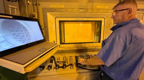 Kate Stephens/BBC News A worker at Sellafield removes used nuclear fuel from canisters from behind lead-lined glass, using a remotely operated robotic arm. The worker, in a blue shirt, stands in front of a thick, lead-lined window operating the robotic arm via a control panel in front of him.