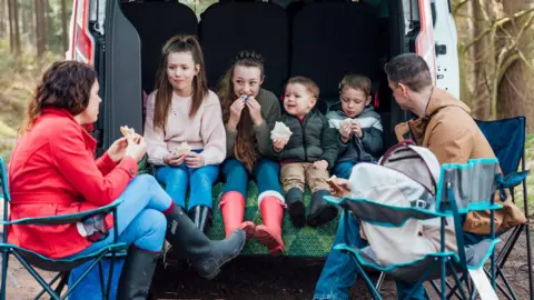 Children sitting together in the boot of a van in the woods eating packed lunch and two adults sitting on camping chairs outside.