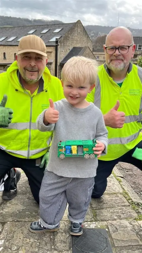 A young boy wearing a grey sweatshirt and holding a green toy truck stands between two waste crew staff, who are also giving the thumbs up. The men are wearing yellow high-visibility jackets