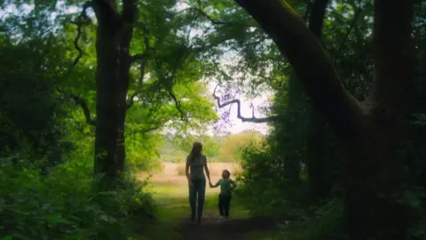 The Witness, Netflix A child riding a small bike on a park path while a woman walks beside him, with trees and grass in the background.