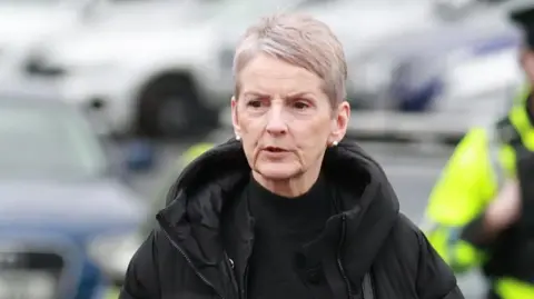 PA Media A woman with short grey hair walking through a carpark wearing a black coat and black high necked top.