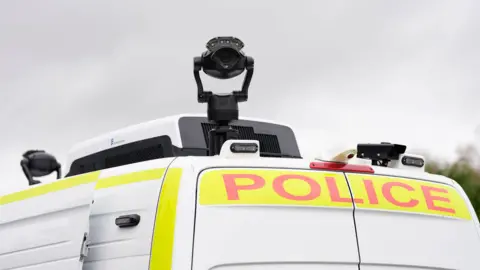 Top of a white police van with a black camera attached on it.