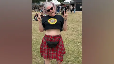 Zoe Durber-Worrall A woman with pink and white hair is wearing black sunglasses, a black top with the yellow and red Guns N' Roses logo on it, and a black tartan skirt with a black bum bag. She is sticking out her tongue and has both hands up in the "rock" signal.
