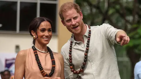 AFP Prince Harry and Meghan, Duchess of Sussex, both smile while wearing large necklaces while Harry points to something out of frame, while in Nigeria in May