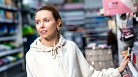 BBC/Little Dooley/Daniel Harrison Stacey Dooley in a beauty shop. A woman with ginger hair and a white hoodie stands in a beauty shop with her hand on a product.